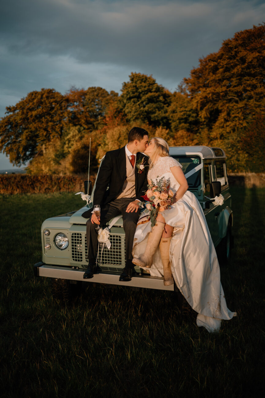 Bride and groom sat on a landrover bonnet kissing, the bride is wearing wellies